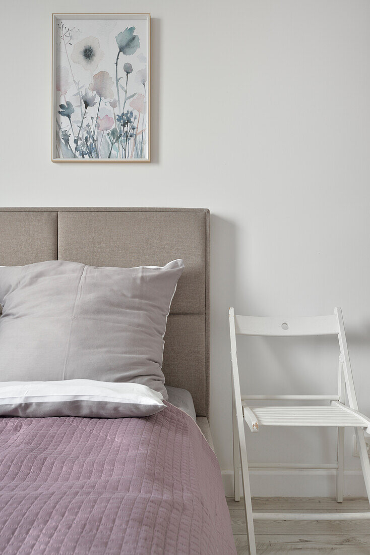 Bed with lilac bedspread and upholstered headboard, white chair and wall art