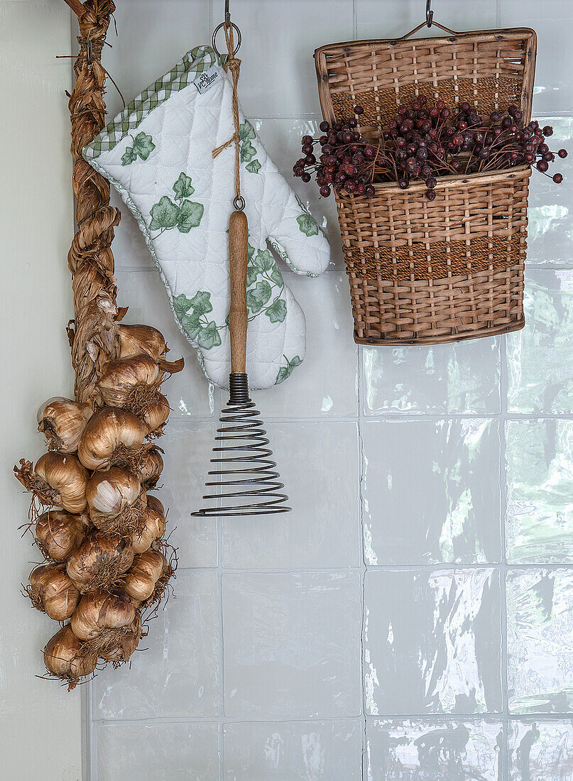 Zopf aus Zwiebeln, Topfhandschuh mit Efeumuster und Korb mit Trockenbeeren an gefliester Wand