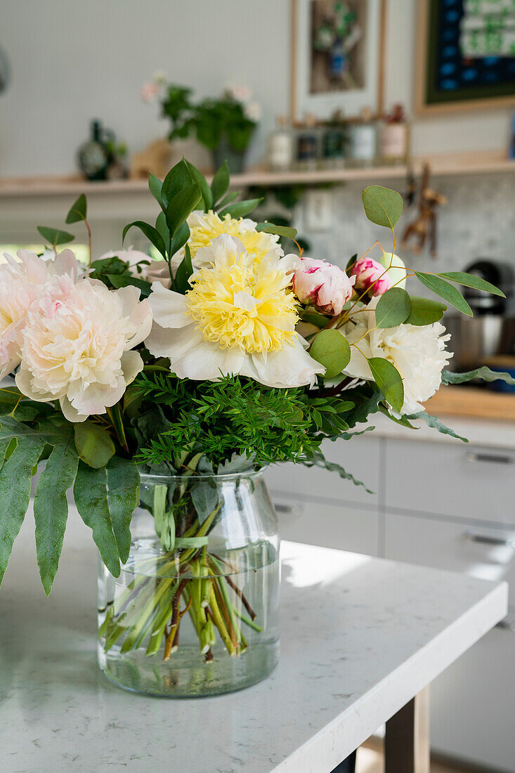 Frischer Blumenstrauß mit Pfingstrosen und Farnen in Glasvase auf Küchenarbeitsplatte