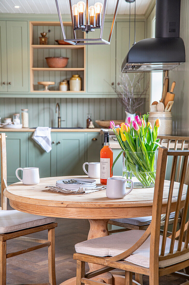 Round wooden table with bouquet of tulips in country-style kitchen with pastel green cabinets