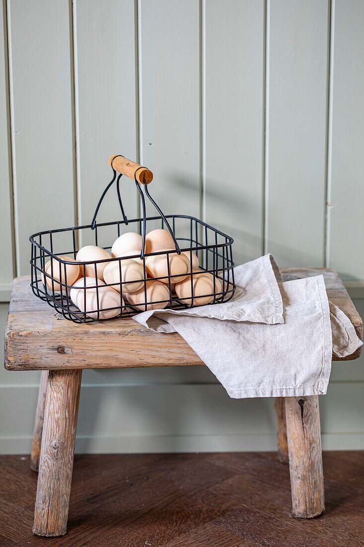 Wire basket with fresh eggs on a rustic wooden stool and tea towel