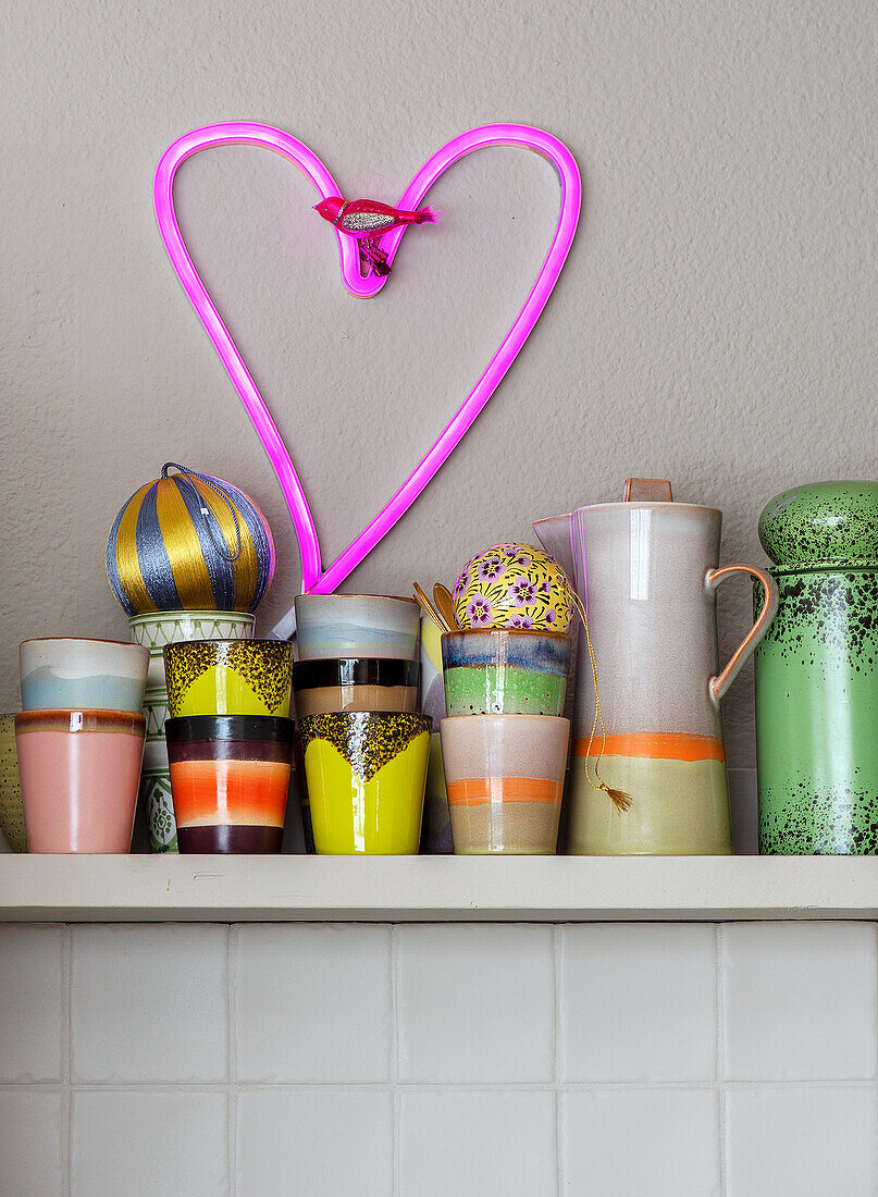 Colourful cups and jugs on shelf and neon-coloured heart as wall decoration