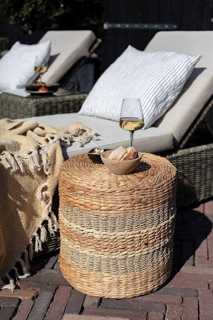 Weißwein und Snacks auf geflochtenem Rattan-Hocker neben Sonnenliegen im Garten