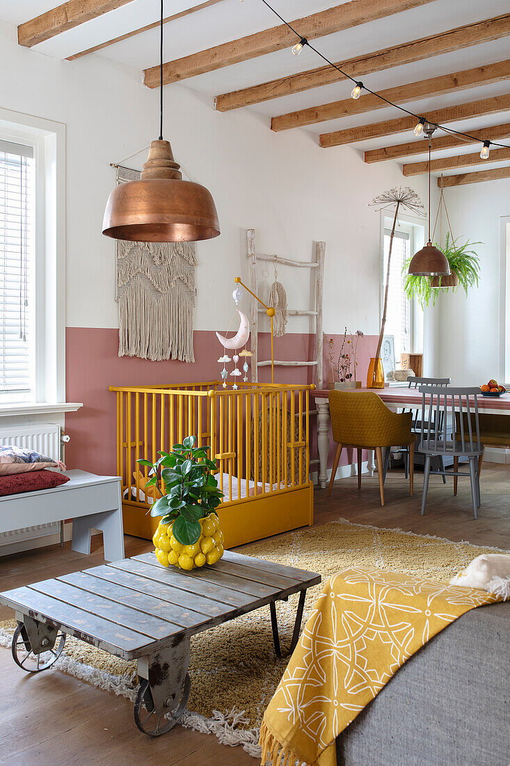 Open-plan living room with cot, macramé wall hanging and wooden beamed ceiling