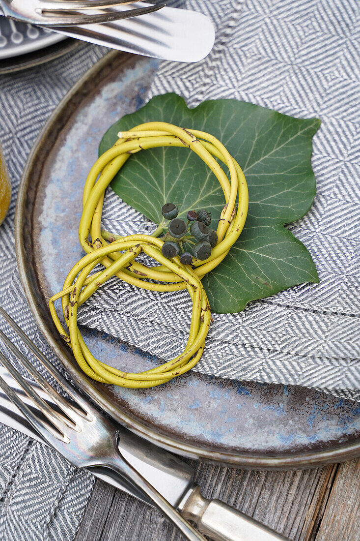 Napkin rings made of twigs on a green leaf and patterned fabric napkin with ivy berries next to silver cutlery