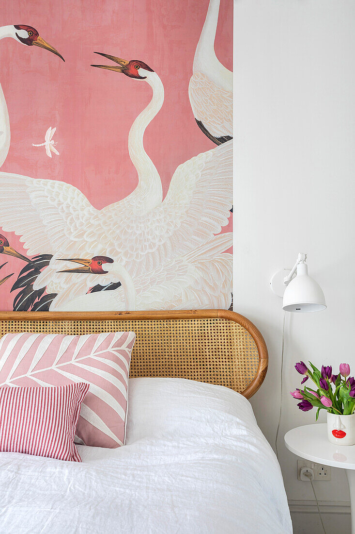 Bed with rattan headboard, pink wallpaper with herons and bouquet of tulips on bedside table in bedroom