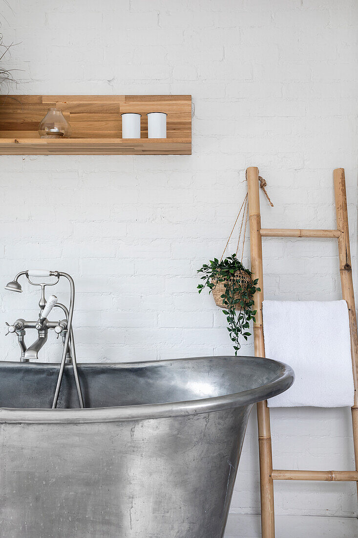 Freestanding metal bathtub in front of white brick wall, bamboo ladder and hanging basket