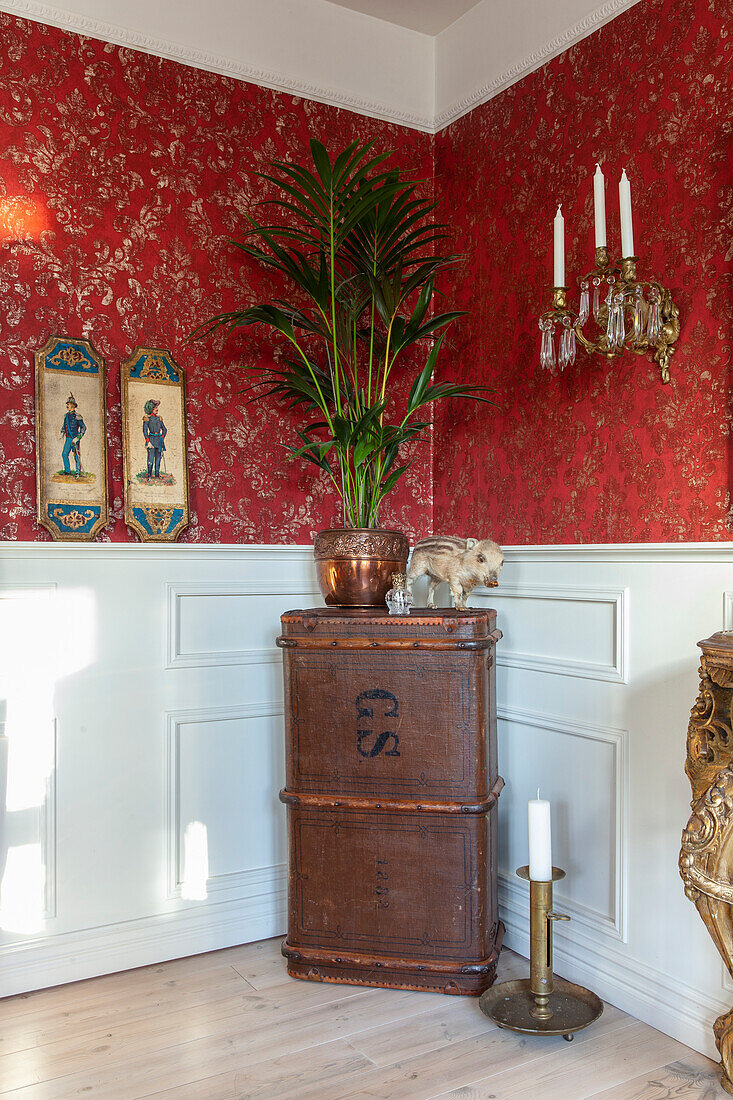 Antique chest with palm tree in a pot and wall candle holder in front of red wallpaper