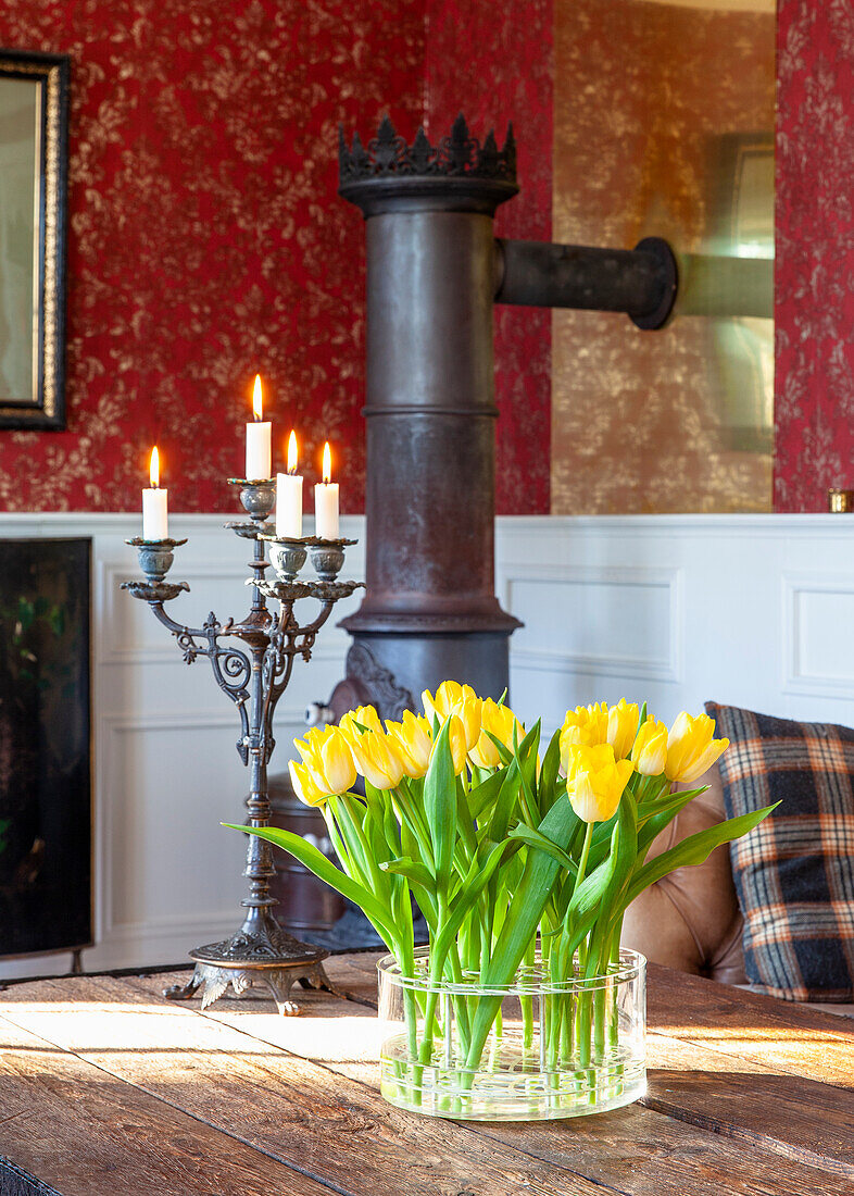 Gelbe Tulpen (Tulipa) in Glasvase auf Holztisch, antiker Kaminofen im Hintergrund