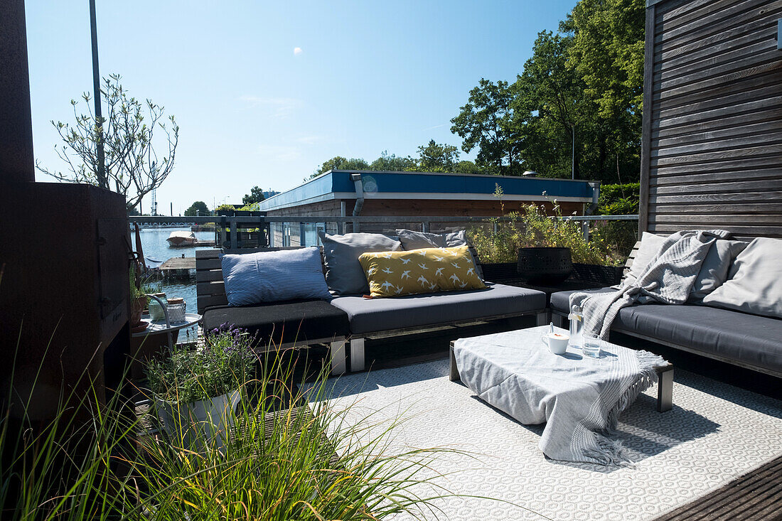 Gartenlounge mit grauen Sofas und Kissen auf Holzterrasse am Flussufer