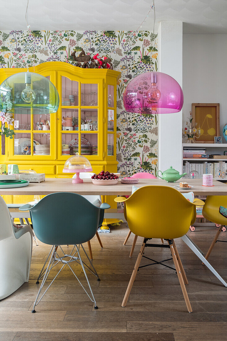 Esszimmer mit bunten Schalenstühlen, floraler Tapete und gelbem Vitrinenschrank