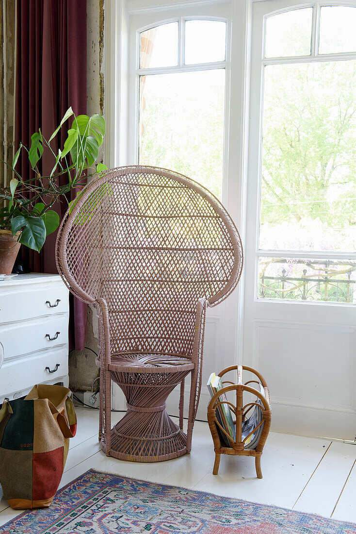 Rattan chair in front of large window in bright room with potted plant and newspaper stand