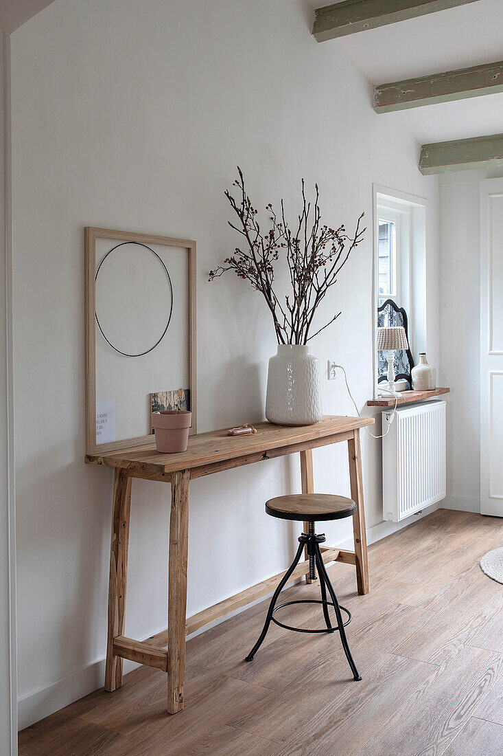 Wooden table with branches in vase and stool in hallway with wooden floor and wall decoration