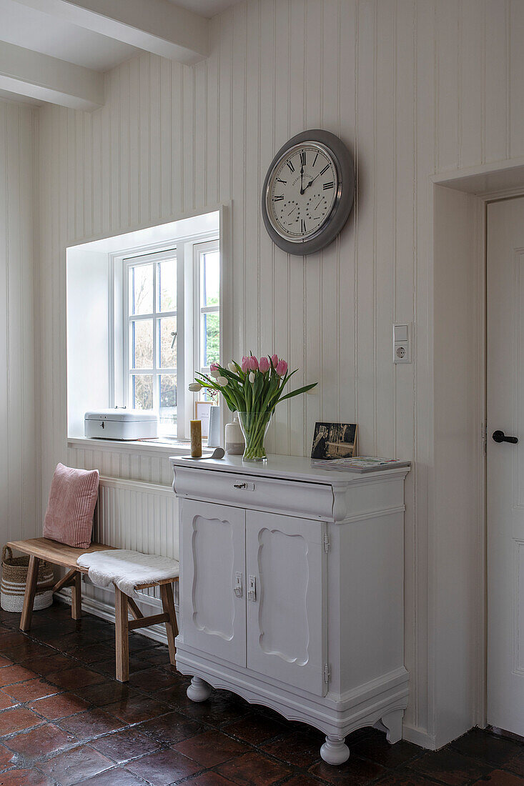 White hall cupboard with bouquet of tulips and wall clock in front of wood panelling