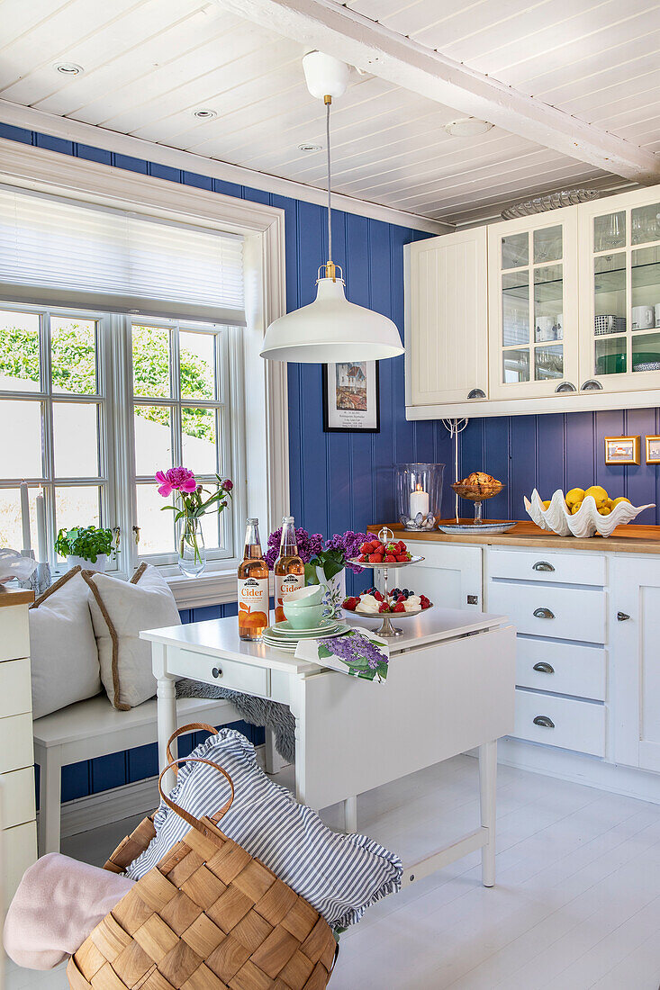 Country-style kitchen with blue walls and white cabinets, folding table in front of window