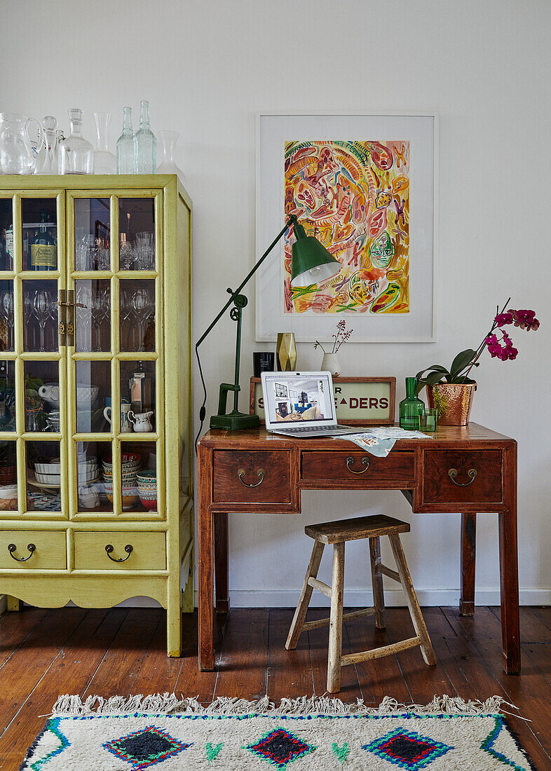 Vintage Schreibtisch mit Hocker vor einer Wand mit abstraktem Kunstwerk und grünem Vitrinenschrank