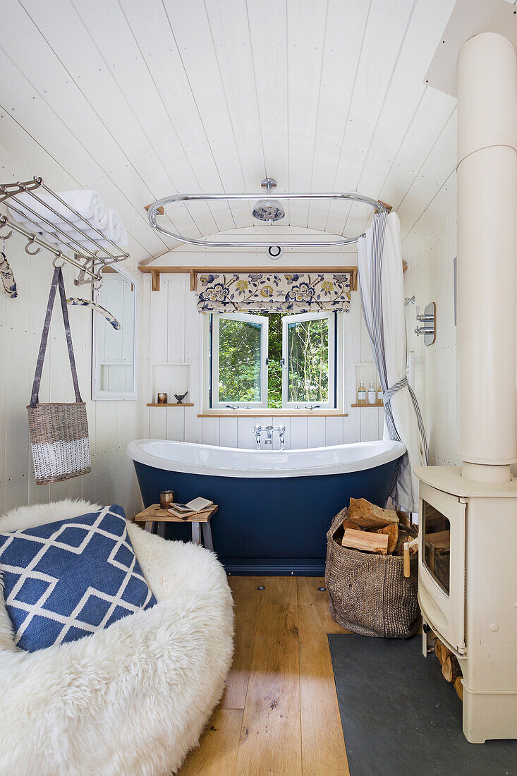 Freestanding bathtub in a small, cosy bathroom with wood-burning stove and sheepskin armchair