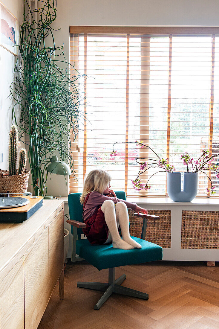 Child on a green swivel chair in front of a window with wooden blinds and plants