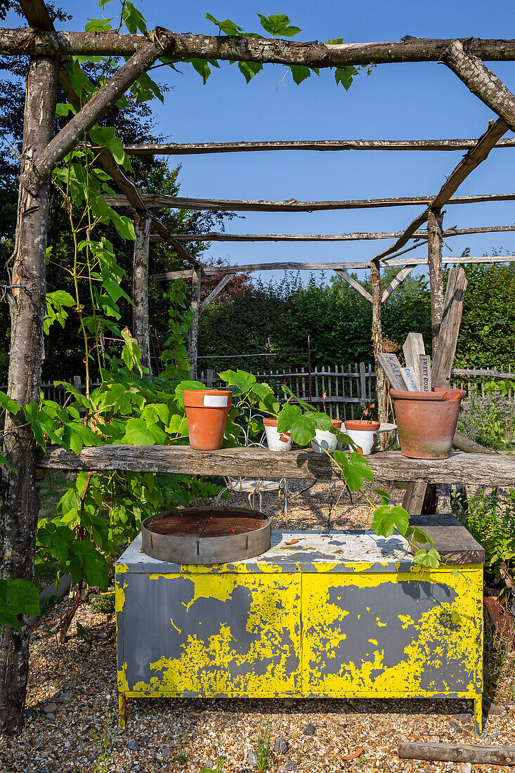 Rustikaler Gartentisch mit abblätternder gelber Farbe und Blumentöpfen unter Pergola