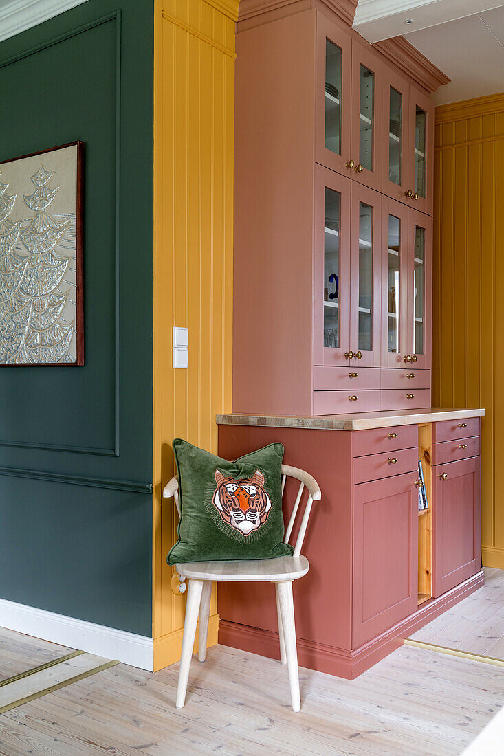 Terracotta-colored built-in cupboard with glass doors and green cushion on wooden chair in front of yellow wall