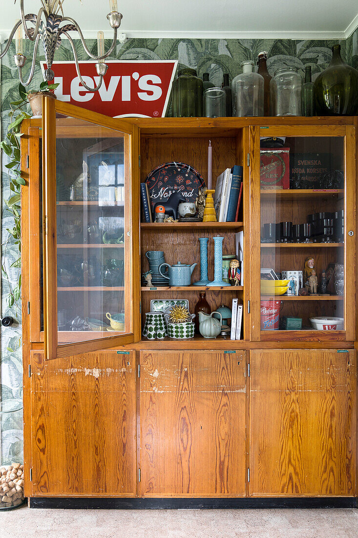 Vintage-Holzschrank mit Glastüren und Retro-Gegenständen vor Tapete mit Palmenmuster