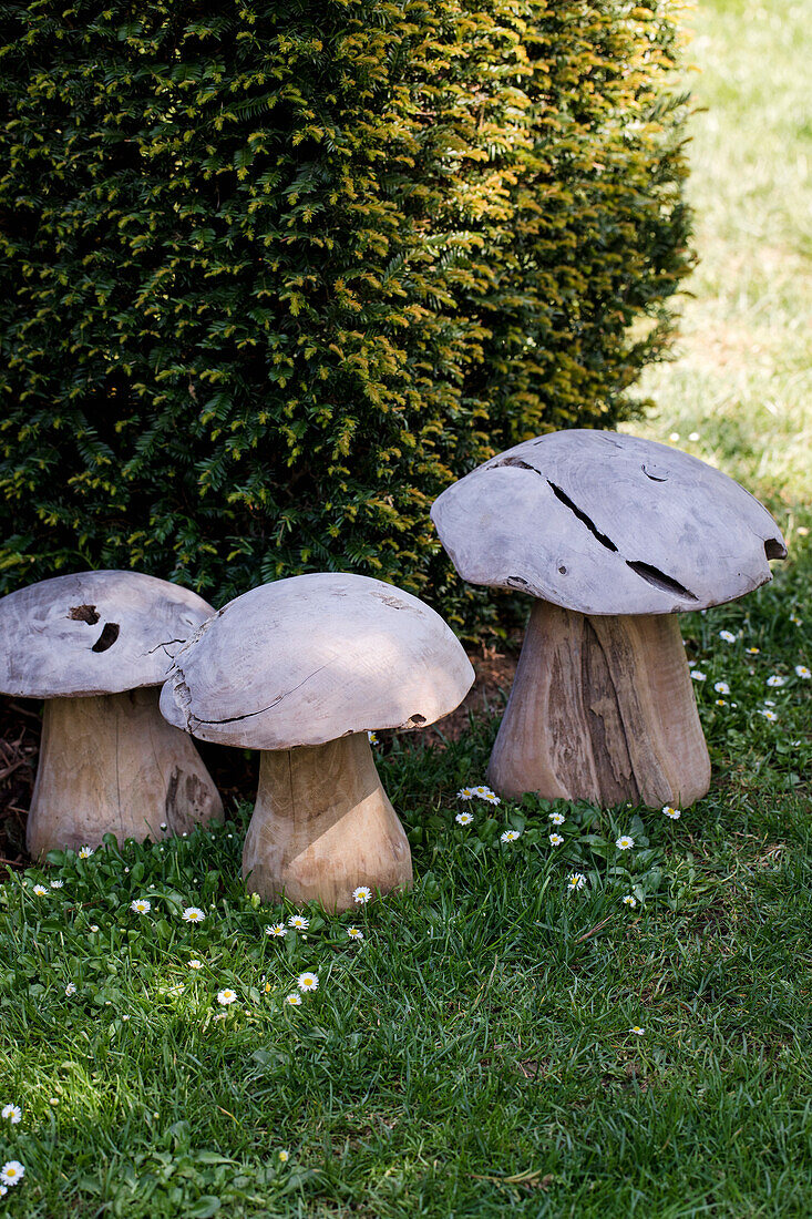 Drei dekorative Pilzskulpturen aus Holz im Garten neben einer Hecke