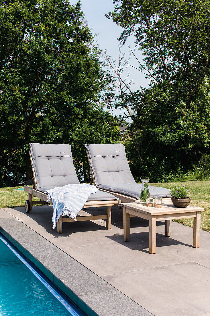 Zwei Liegestühle mit grauen Polstern am Pool, Beistelltisch aus Holz