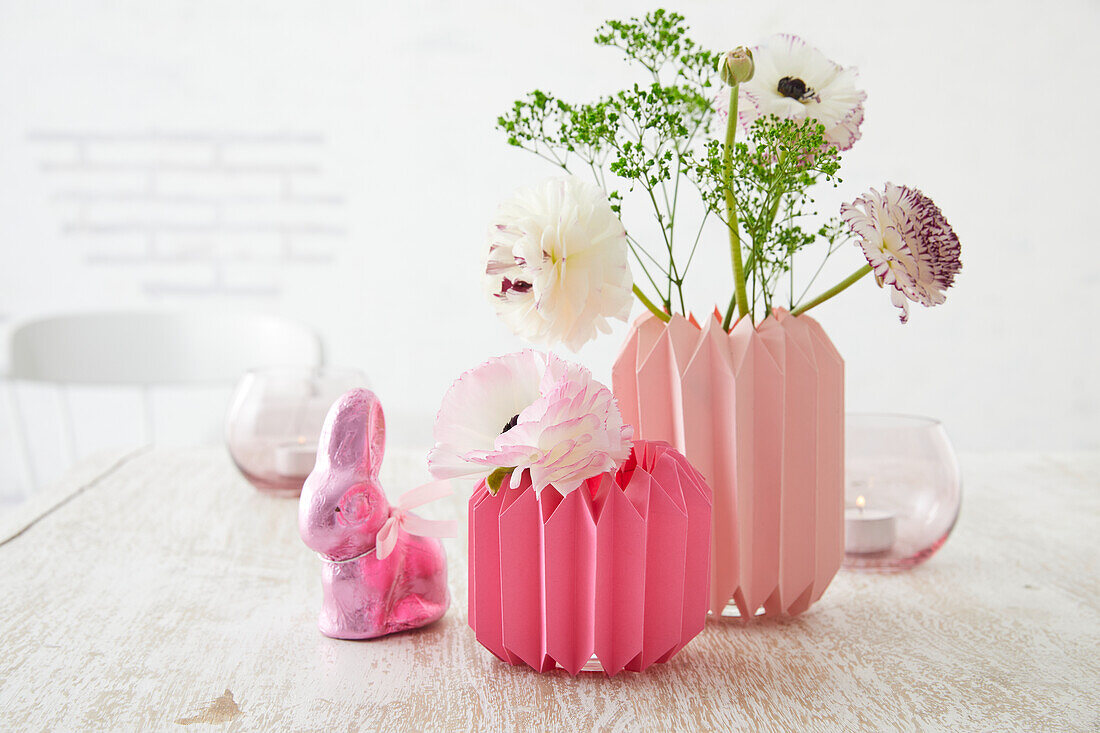 Homemade vases made from folded paper with spring flowers on a light-colored wooden table