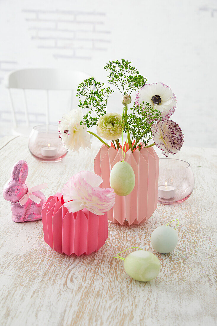 Selbstgemachte Vasen aus gefaltetem Papier mit Frühlingsblumen, Dekohasen und Ostereier auf Holztisch