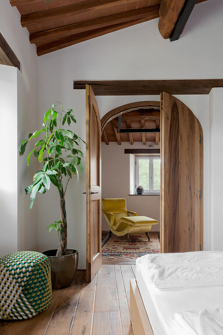 Schlafzimmer mit Holzboden, Pouf und großer Pflanze, Blick ins angrenzende Zimmer