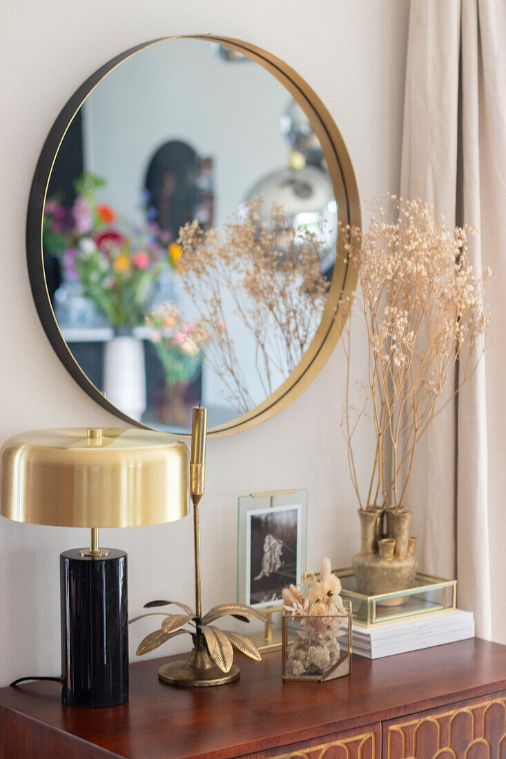 Runder Spiegel mit goldener Umrandung über dekoriertem Sideboard mit Trockenblumen und goldener Tischlampe