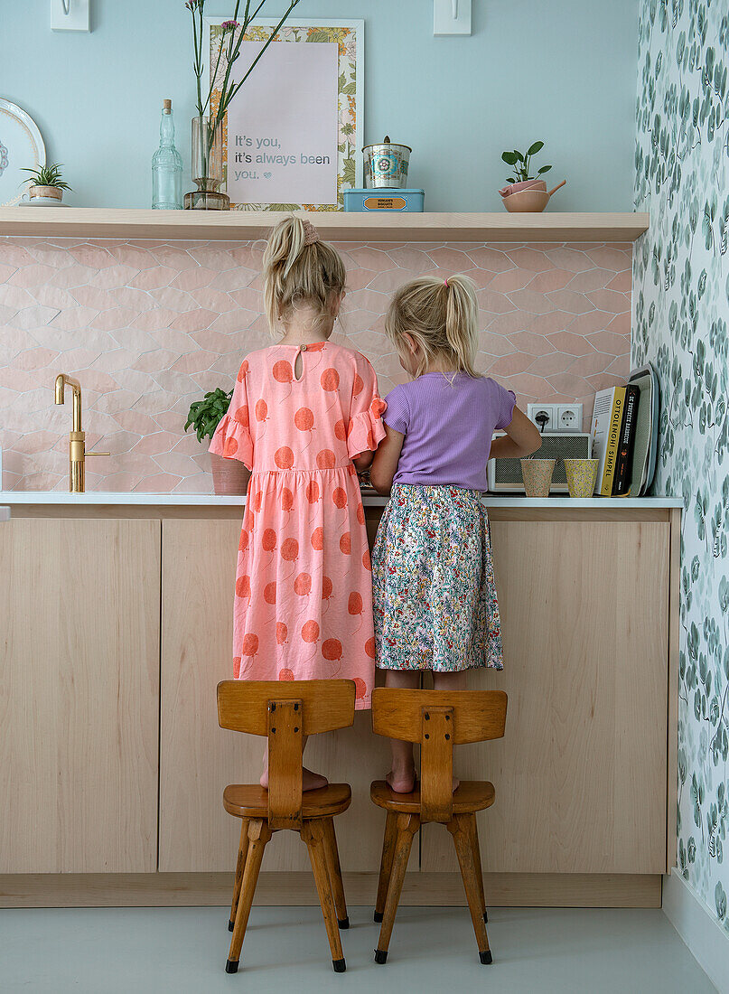 Two children on chairs in a pastel-colored kitchen with floral wallpaper