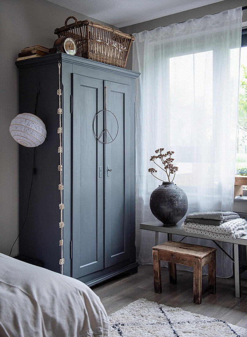 Grey-painted wardrobe with basket in bedroom, decorative vase on small table next to it