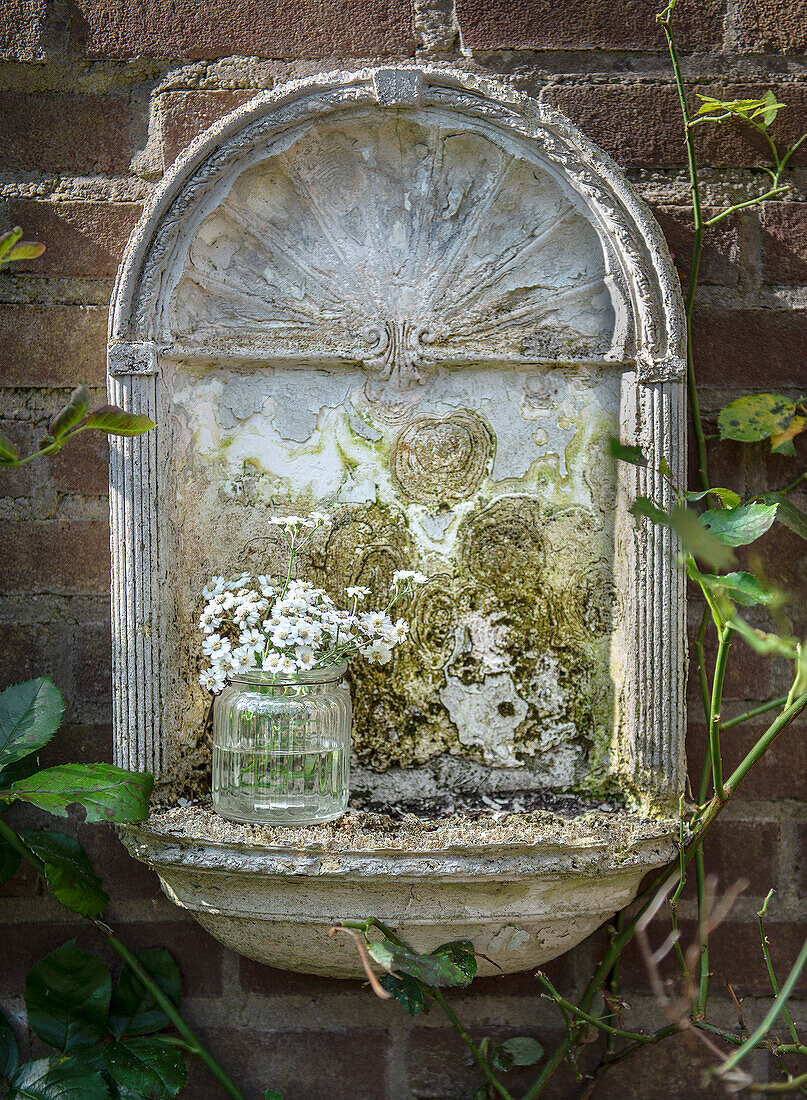 Antiker, stillgelegter Wandbrunnen mit Glasvase und weißen Blumen