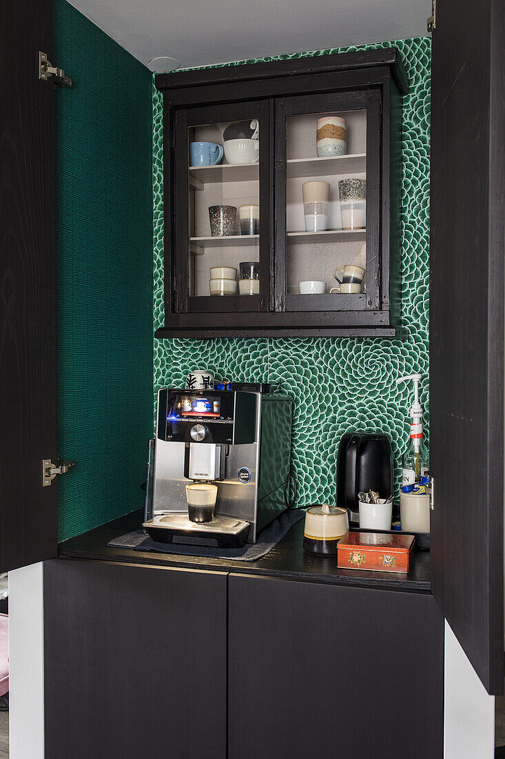 Coffee machine on a black worktop with display cabinet and green patterned wallpaper