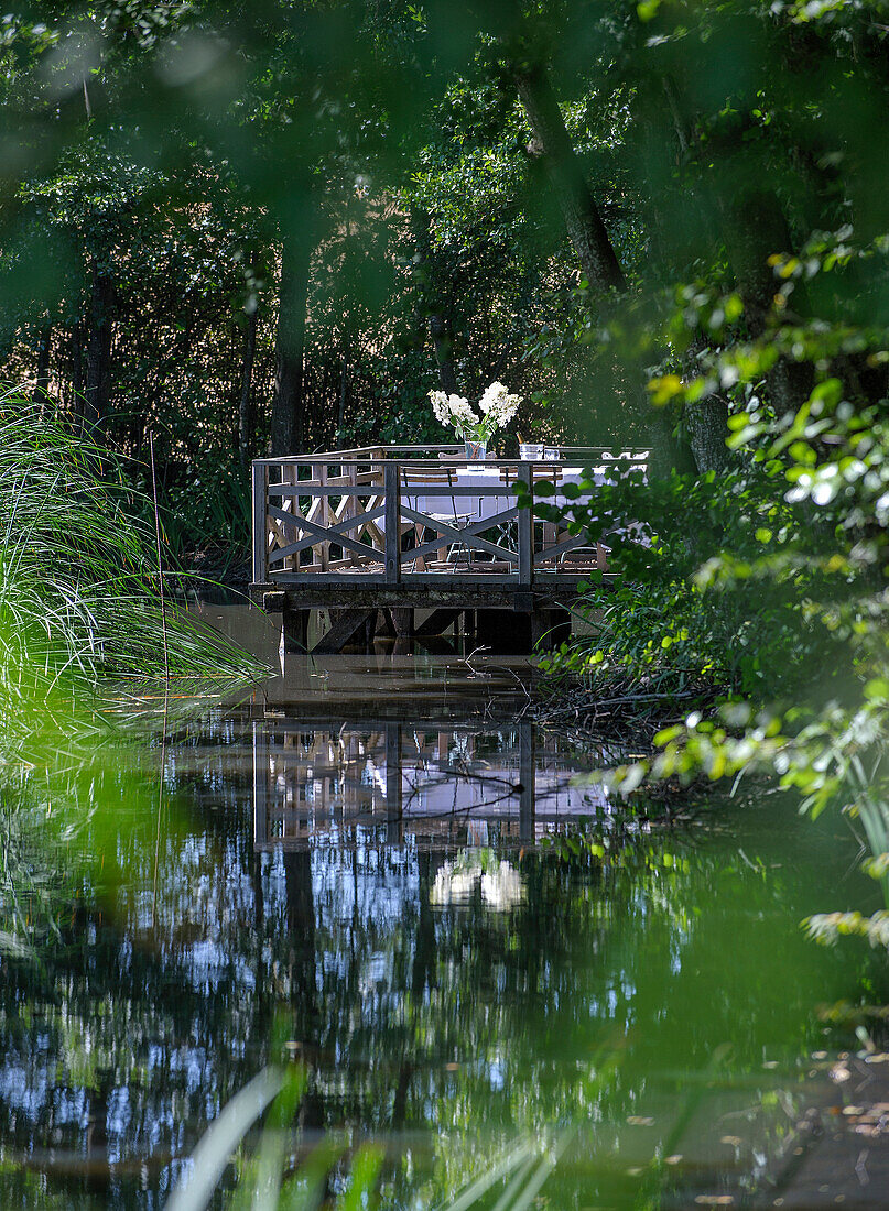 Holzterrasse mit Tisch und Stühlen am Ufer eines ruhigen Teichs im Garten