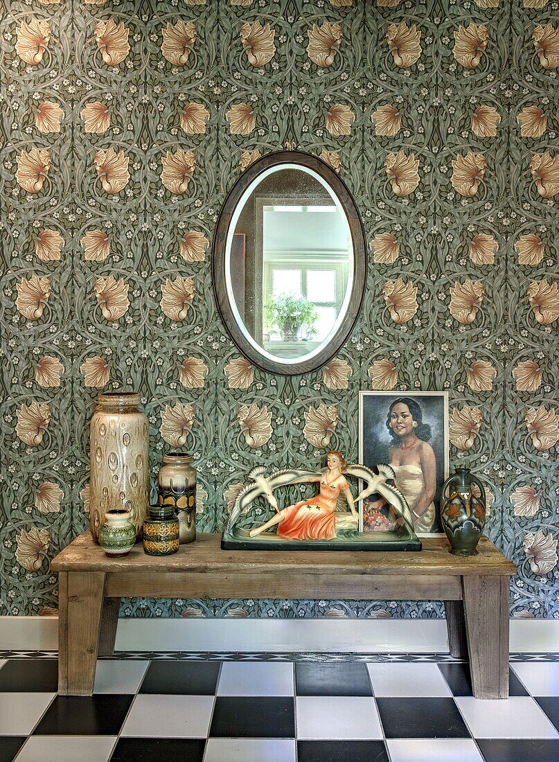 Vases, figurine and picture on wooden bench in front of patterned wallpaper in hallway