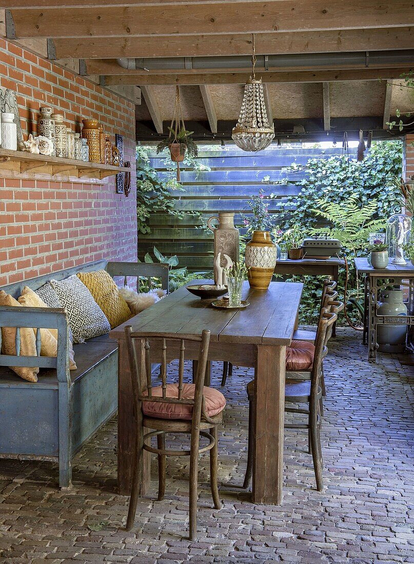 Rustic dining area under a covered terrace with wooden table, old chairs and brick wall