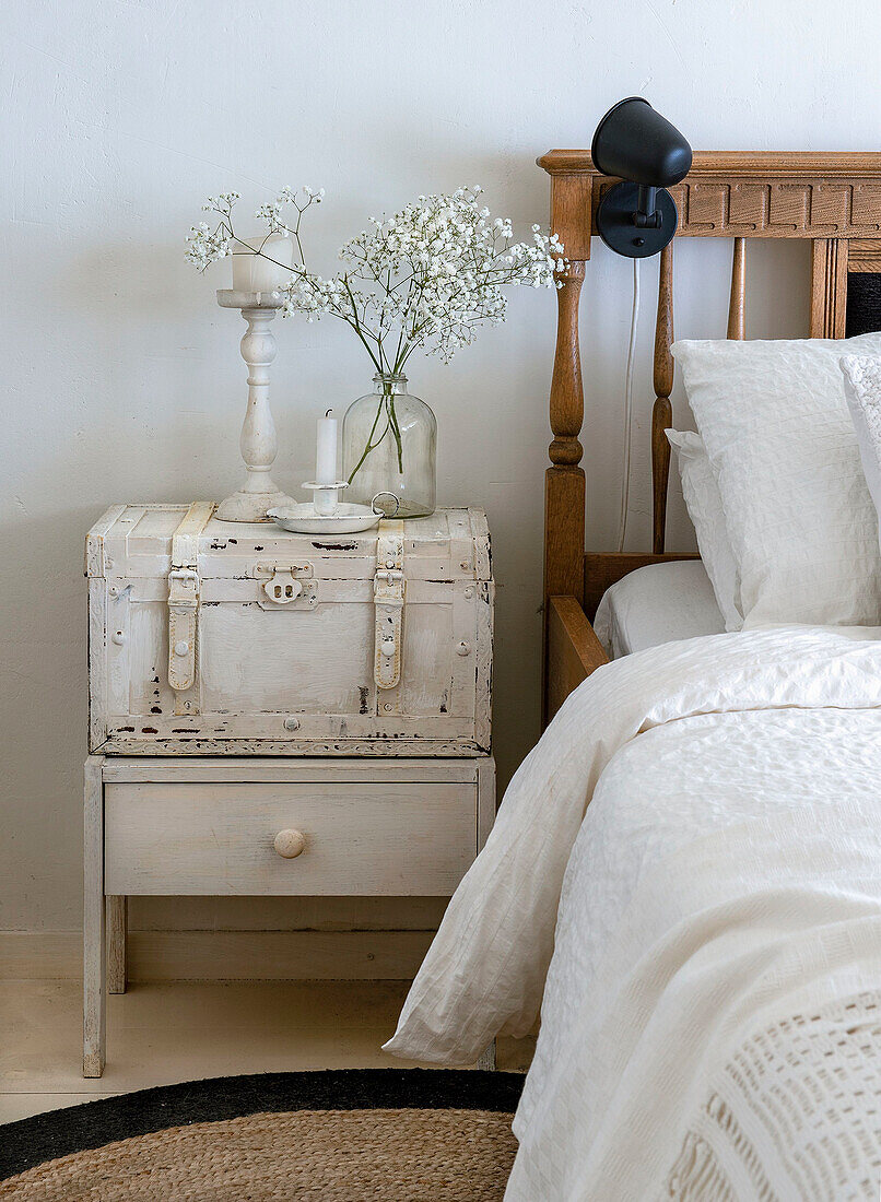 Holz-Nachttisch mit Vintage-Koffer und Vase mit Schleierkraut neben einem rustikalen Bett