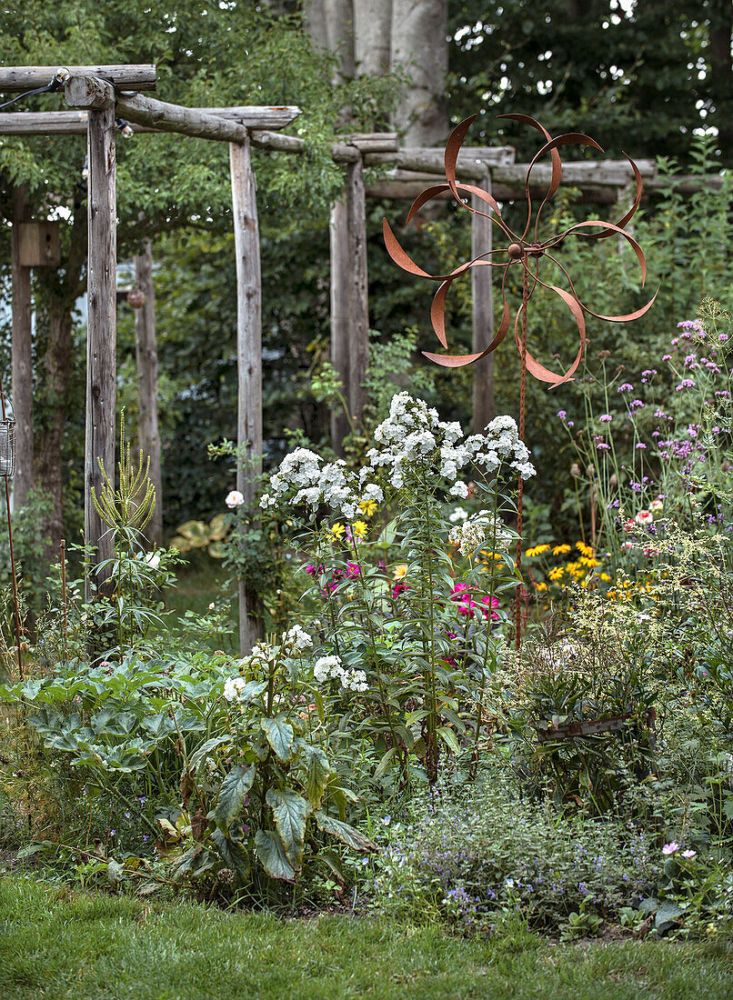 Blumenbeet mit Metallwindrad und Holzspalier im Garten