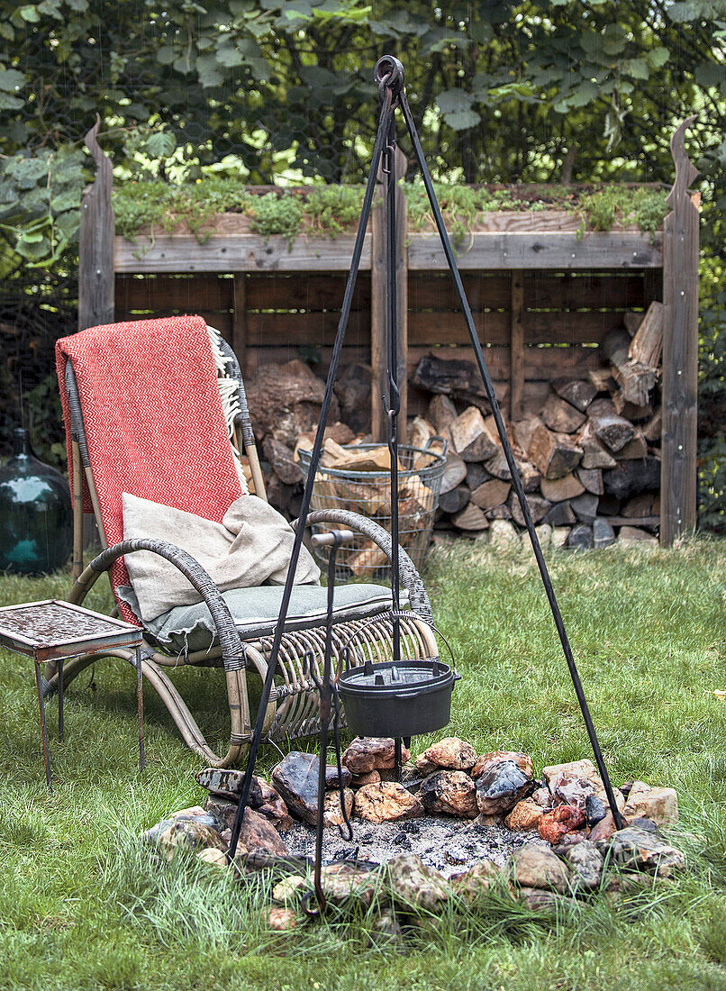 Feuerstelle mit Hängekessel und Gartenstuhl vor Holzlager im Garten