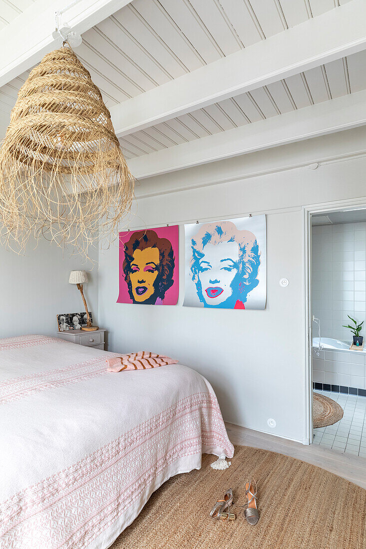 Schlafzimmer mit Rattan-Deckenlampe, Pop-Art-Bildern und Bett mit rosafarbener Tagesdecke