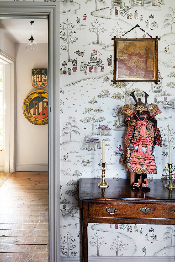 Console table with antique candle holders and traditional doll in front of wallpaper with landscape motifs