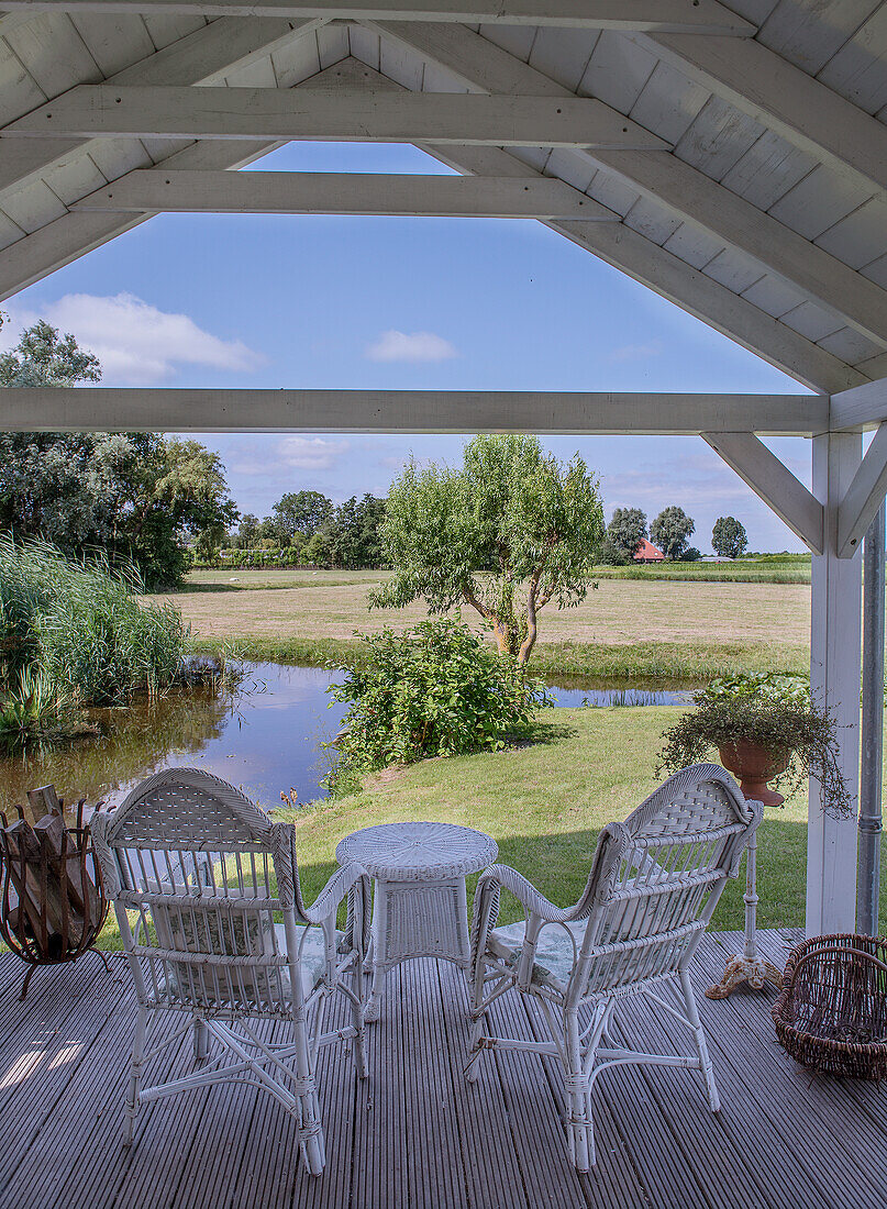 White rattan armchairs and table under a covered terrace with a view of a pond