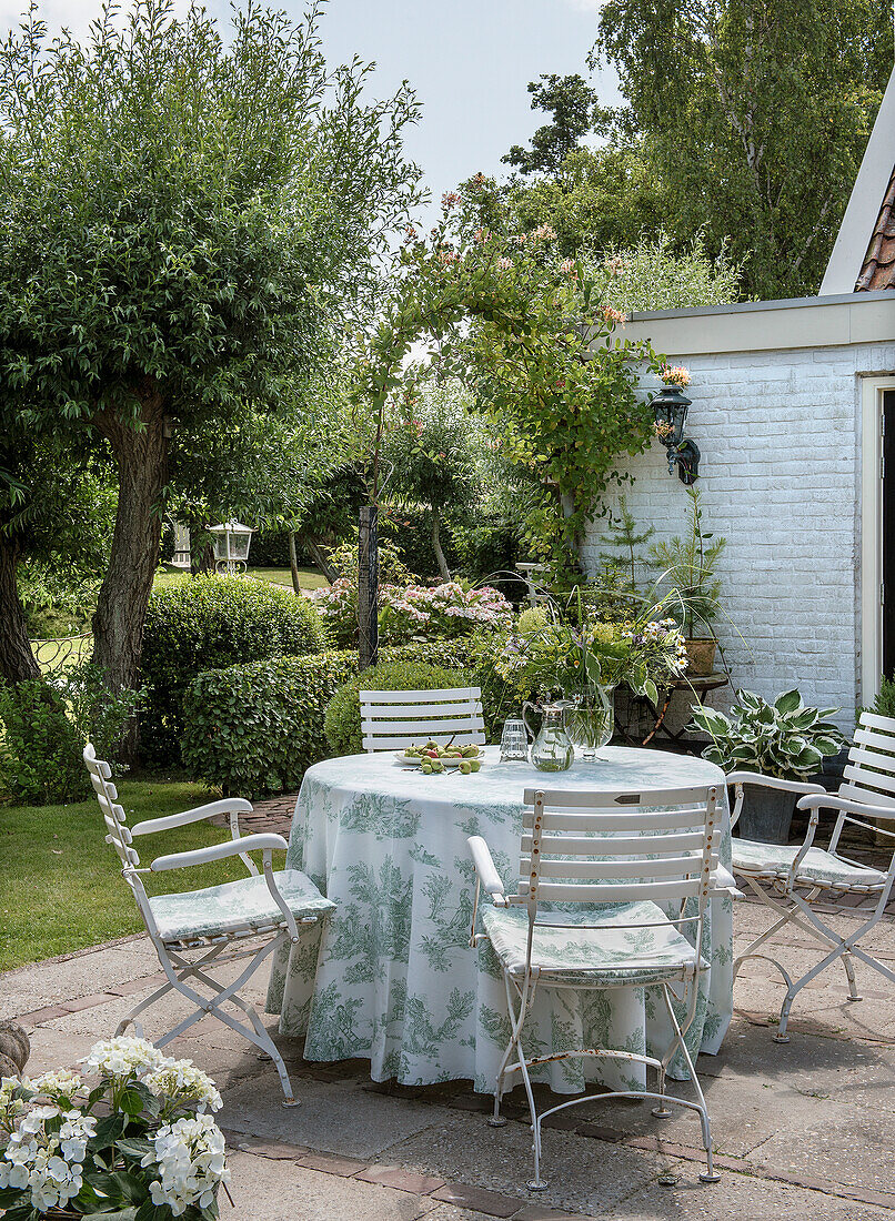 Runder Gartentisch mit weiß-grüner Tischdecke und Gartenstühlen im Sommergarten