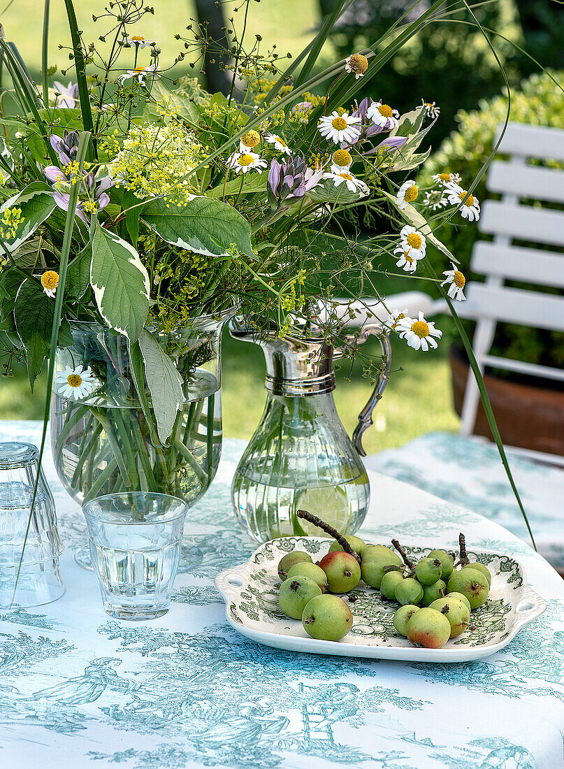 Glasvase und Karaffe mit Wiesenblumen und Teller mit Äpfeln auf einem gedeckten Gartentisch