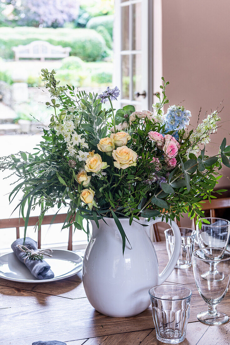 Blumenstrauß in weißer Vase auf gedecktem Holztisch vor offenem Fenster zur Terrasse