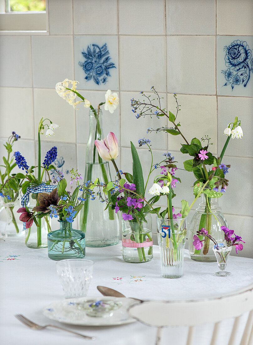 Frühlingsblumen in verschiedenen Glasvasen auf einem weißen Tisch vor Fliesenspiegel mit Blumenmuster