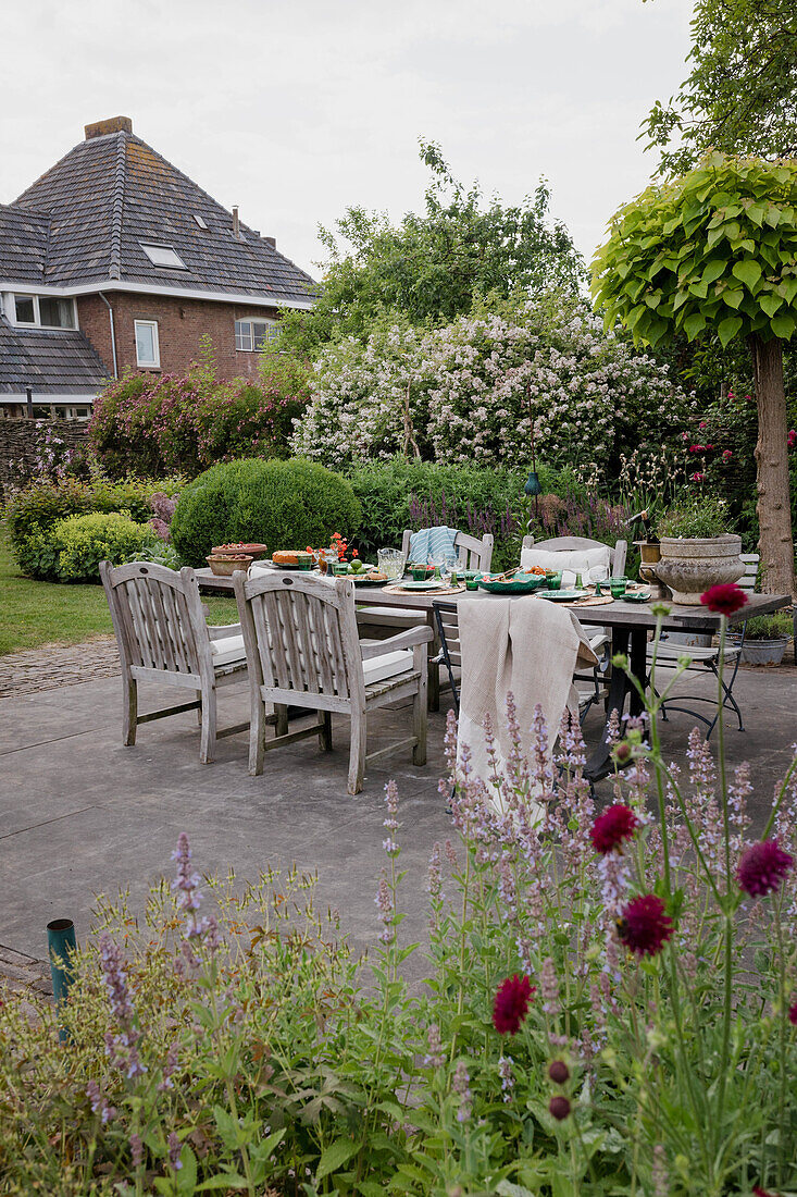 Gedeckter Gartentisch mit Holzstühlen und blühenden Sträuchern im Hintergrund