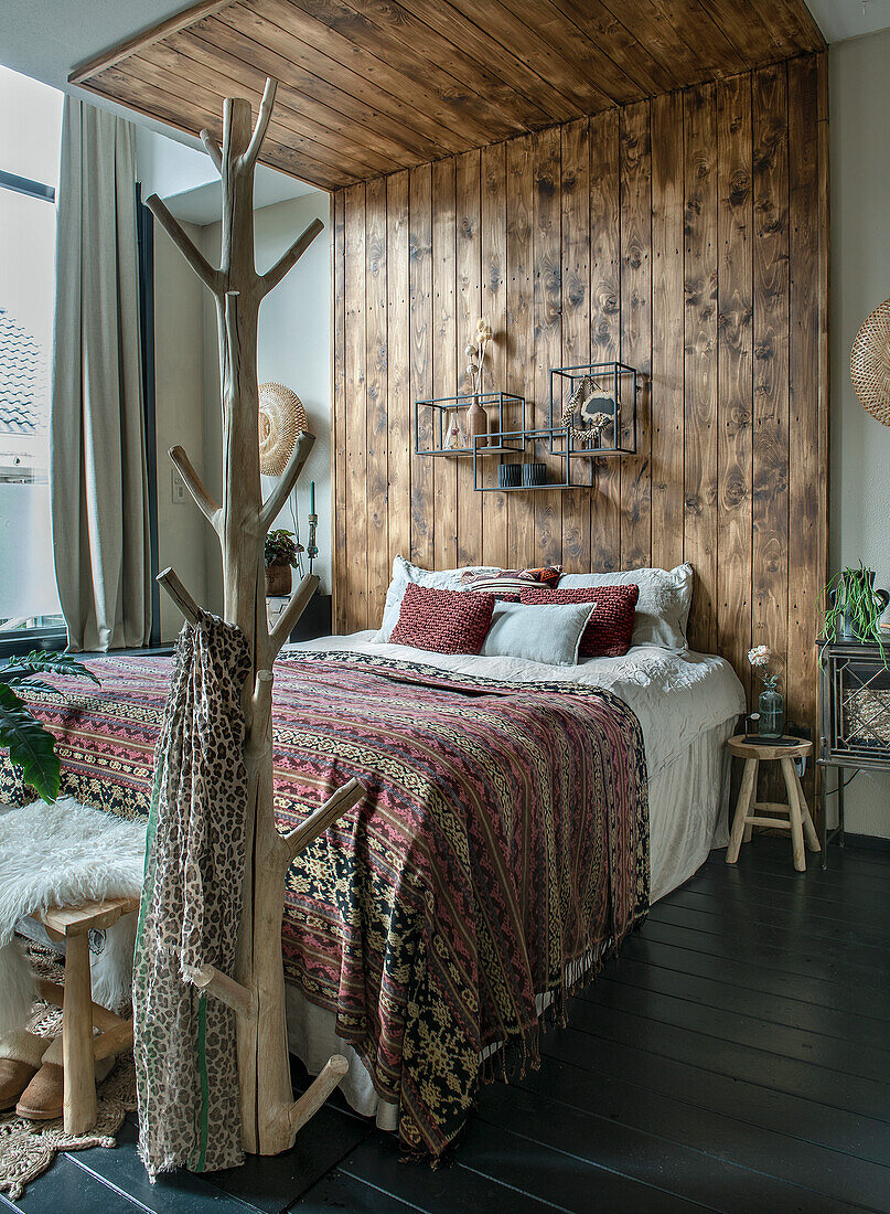 Bed with patterned bedspread in bedroom with wooden wall and clothes rack made of natural wood